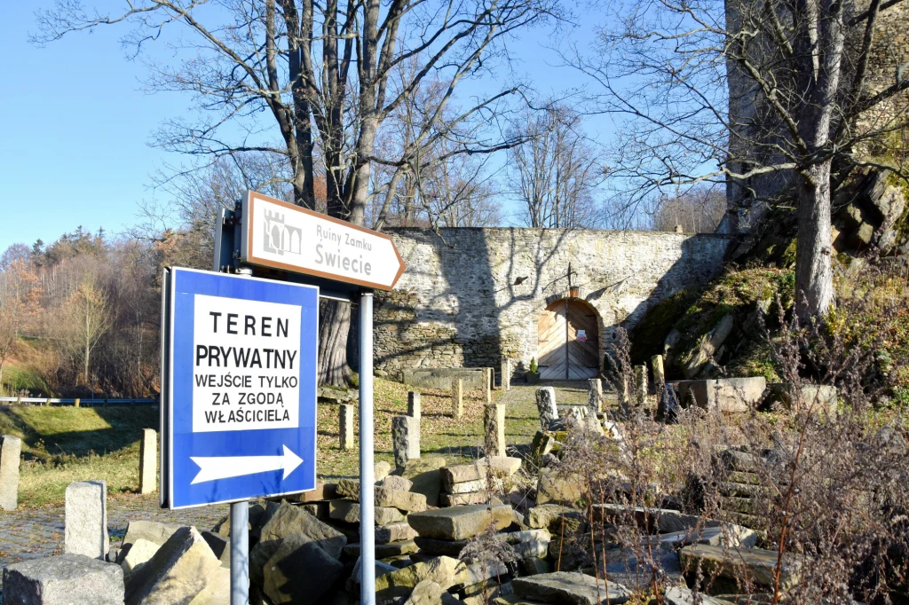 Zamek możemy zwiedzić za 10 zł Wejście na teren zamku Świecie oznaczone tablicą informującą o terenie prywatnym, fragment murów zamku oraz kamienne ogrodzenie, w tle nagie drzewa i słoneczna pogoda.
