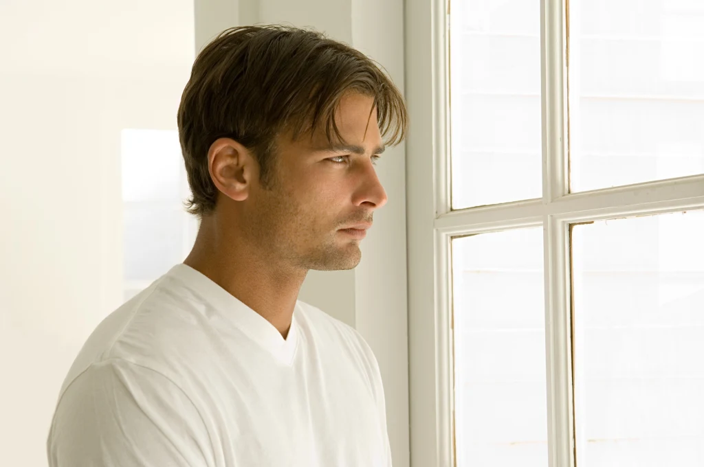 Coraz więcej mężczyzn cierpi na kompleks Adonisa. Czym jest to zjawisko i jak je rozpoznać? Mężczyzna o jasnobrązowych włosach w białej koszulce patrzy przez okno na jasnym tle, z wyrazem zadumy na twarzy.