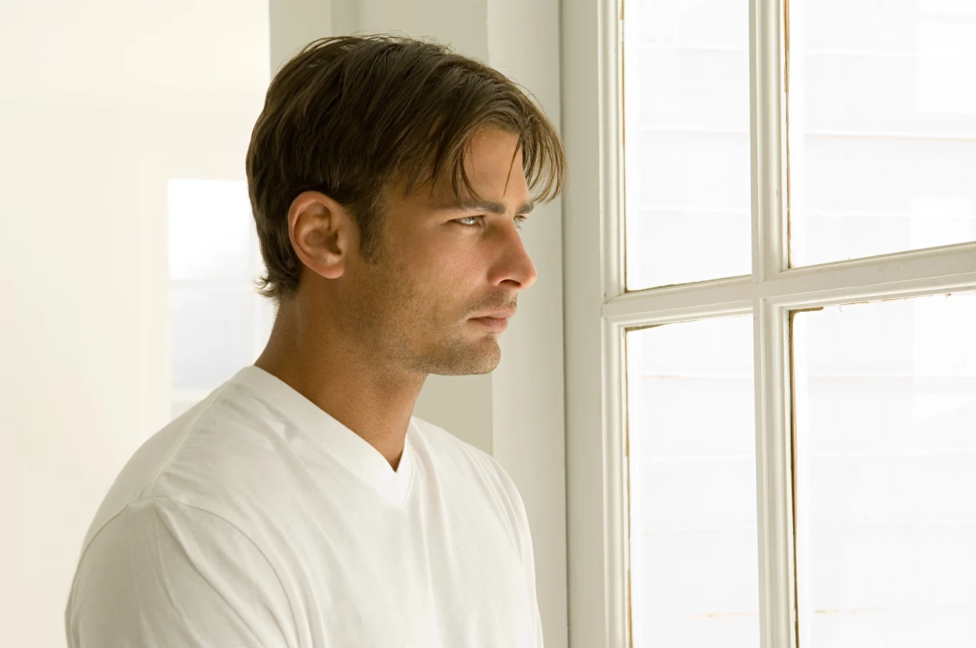Coraz więcej mężczyzn cierpi na kompleks Adonisa. Czym jest to zjawisko i jak je rozpoznać? Mężczyzna o jasnobrązowych włosach w białej koszulce patrzy przez okno na jasnym tle, z wyrazem zadumy na twarzy.