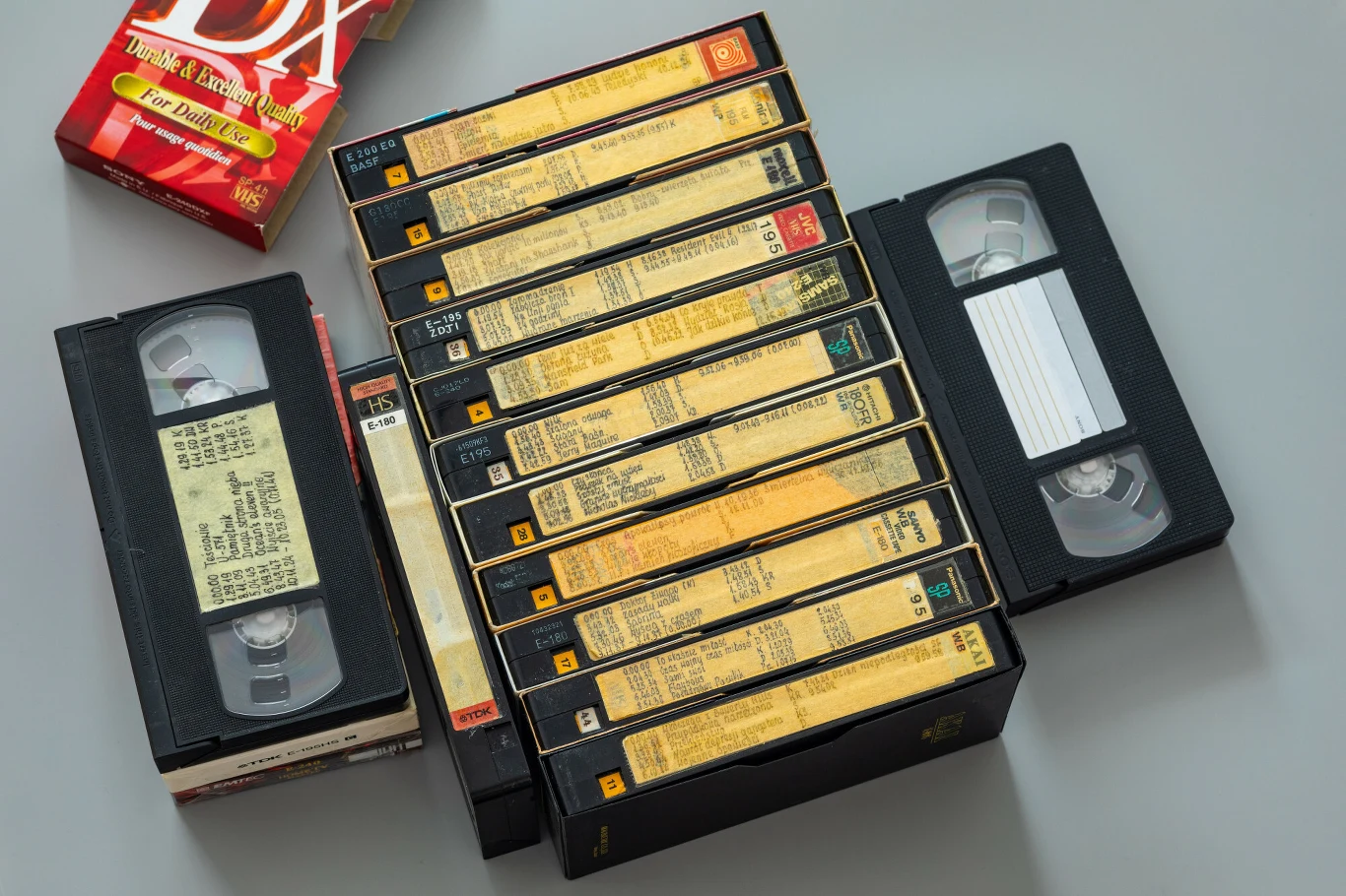 Najrzadsze kasety VHS osiągają wysokie ceny (zdj. ilustracyjne) Kilkanaście kaset wideo VHS ułożonych obok siebie na jasnej powierzchni, z widocznymi etykietami oraz opakowanie po taśmie magnetofonowej w rogu kadru.