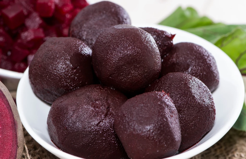 Ugotowane buraki ułożone w białej misce, w tle widoczne pokrojone buraki i zielone liście.