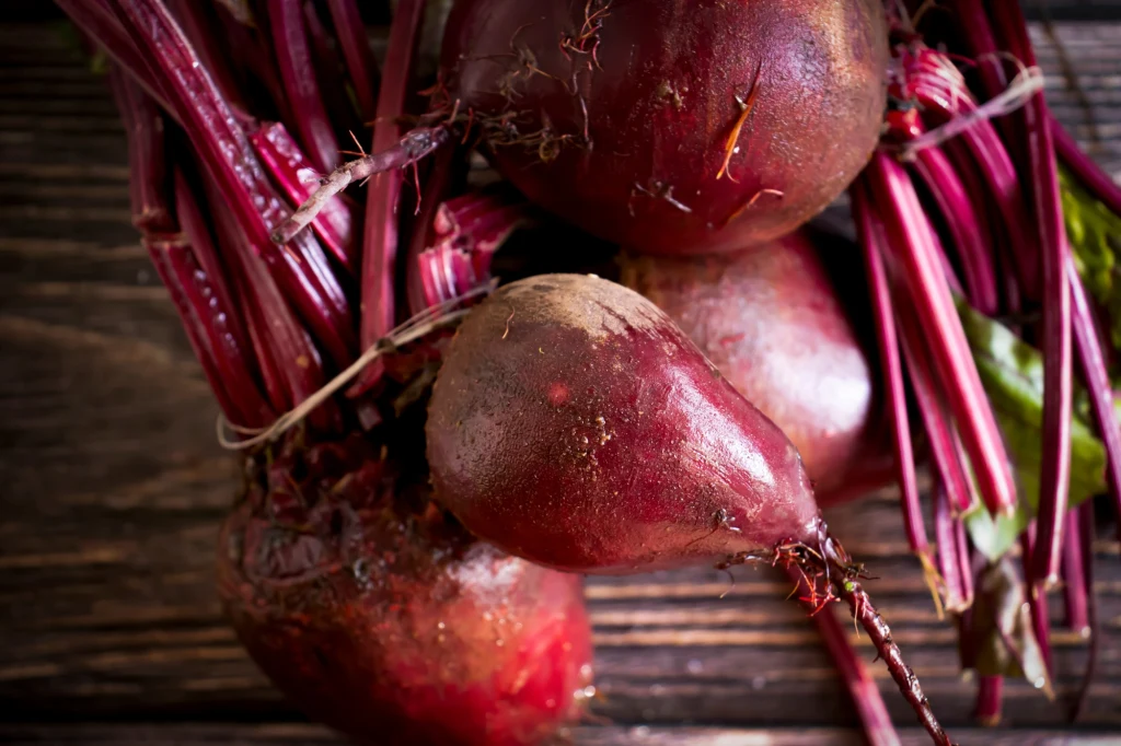 Świeże buraki z liśćmi ułożone na drewnianym stole, ich czerwono-fioletowe korzenie i łodygi kontrastują z ciemnym tłem