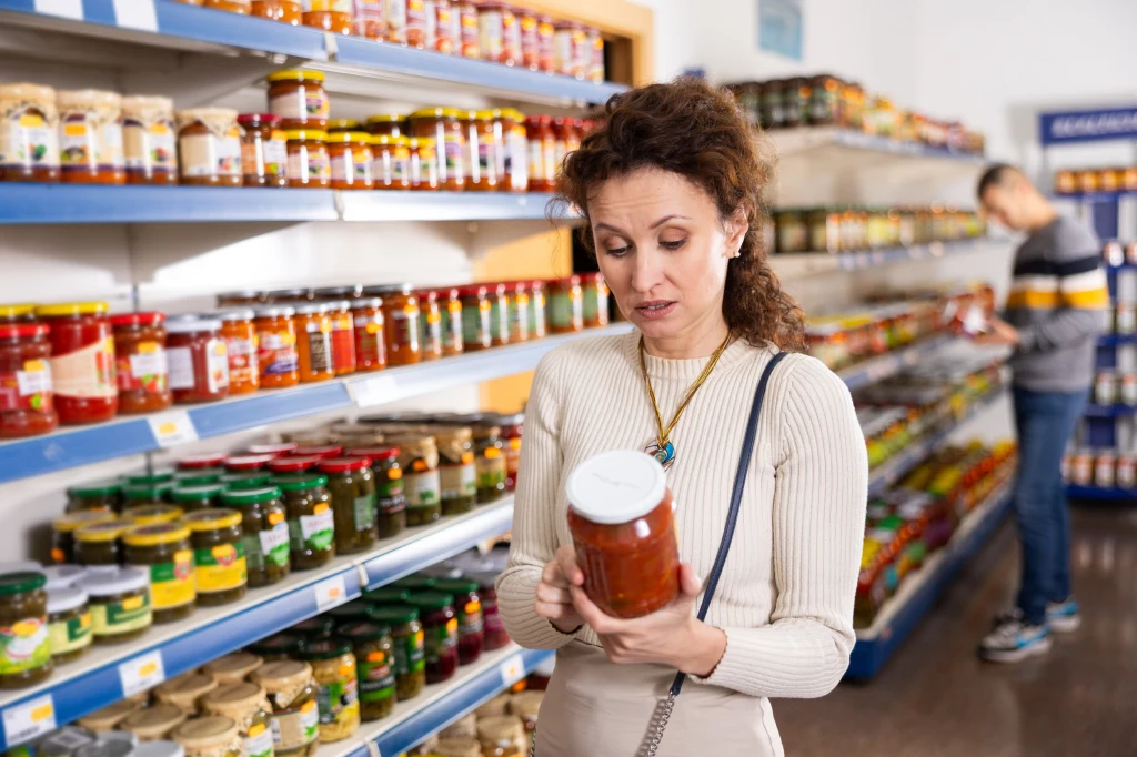Kobieta w jasnym swetrze stoi w alejce sklepu spożywczego, uważnie przyglądając się etykiecie na słoiku, który trzyma w dłoni. Na półkach dookoła znajduje się szeroki wybór przetworów w słoikach. W tle widać jeszcze jedną osobę, przeglądającą produkty.
