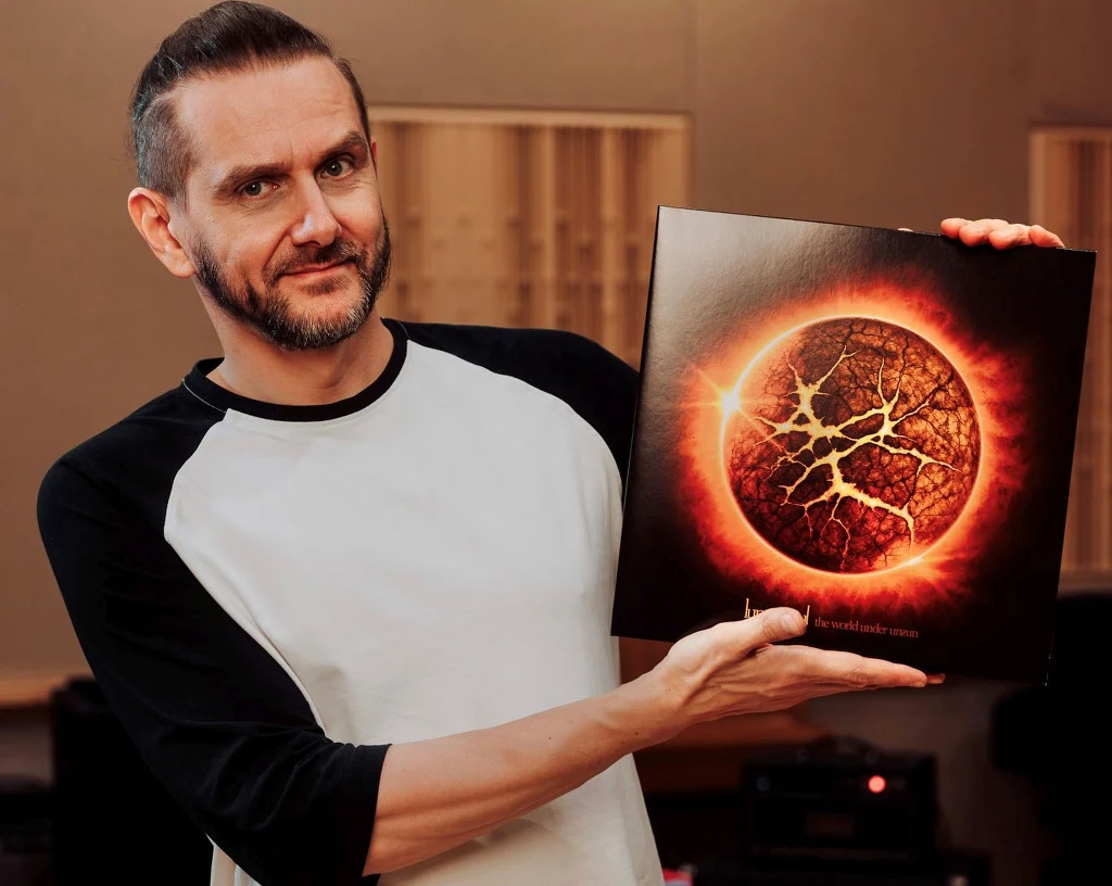 Mężczyzna z zarostem ubrany w biało-czarną koszulkę prezentuje okładkę albumu z motywem płonącej, popękanej planety w intensywnych odcieniach czerwieni i pomarańczu, stojąc w pomieszczeniu ze sprzętem nagraniowym w tle.