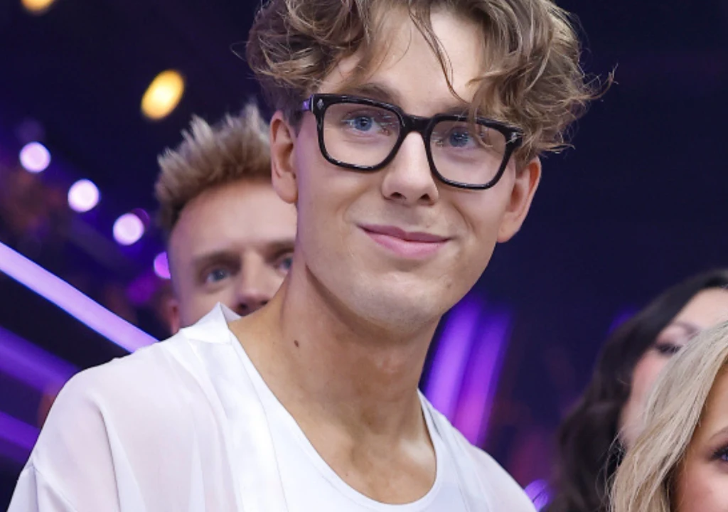 Mikołaj "Bagi" Bagiński Młody mężczyzna w okularach o jasnych, kręconych włosach uśmiecha się lekko, mając na sobie jasną koszulę, w tle kilka osób i kolorowe światła kojarzące się z wydarzeniem rozrywkowym.