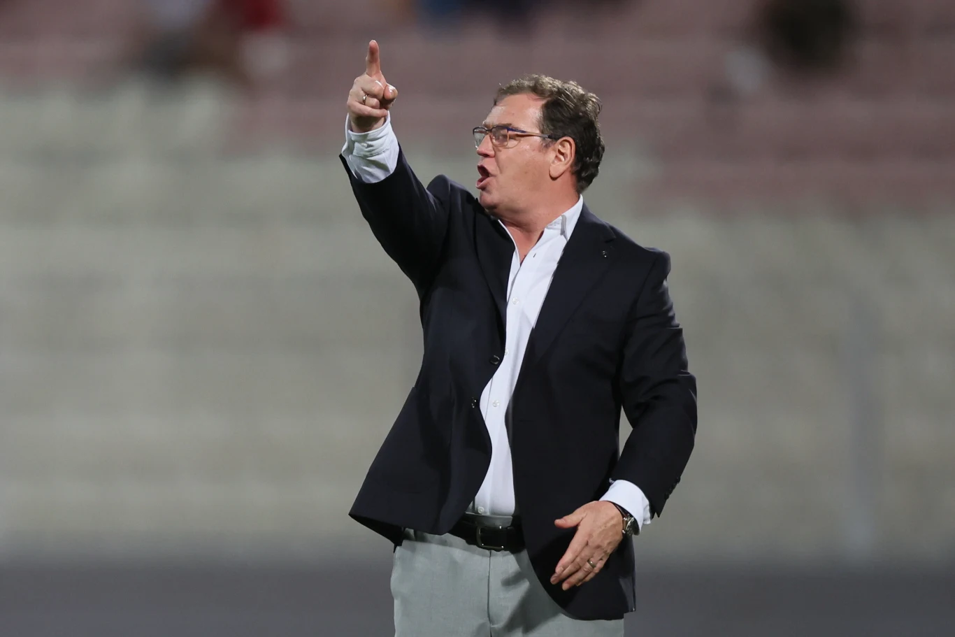 Mężczyzna w średnim wieku w okularach i garniturze energicznie gestykuluje podczas wydarzenia sportowego, stojąc na tle zamazanych trybun.