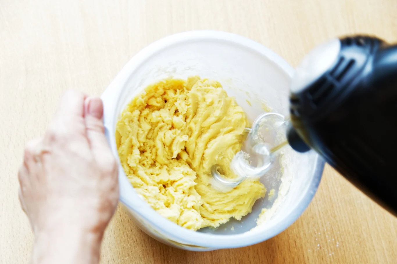 Ręka trzymająca plastikową miskę z żółtą masą, która jest mieszana za pomocą elektrycznego miksera ręcznego, wszystko na jasnym, drewnianym blacie.