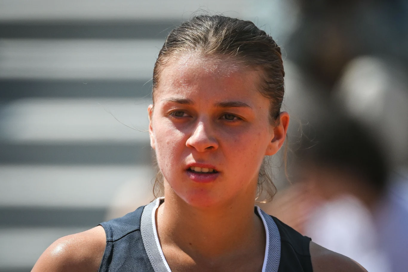 Młoda kobieta o spiętych włosach w sportowym stroju na tle rozmazanego obszaru, prawdopodobnie po wysiłku fizycznym, z lekko spoconą twarzą i skupionym wyrazem.