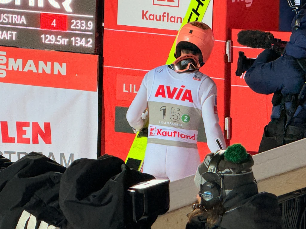 Zawodnik w kombinezonie narciarskim z nartami, stojący tyłem do obiektywu na tle reklam i tablicy wyników, wokół niego znajduje się ekipa medialna z kamerami.