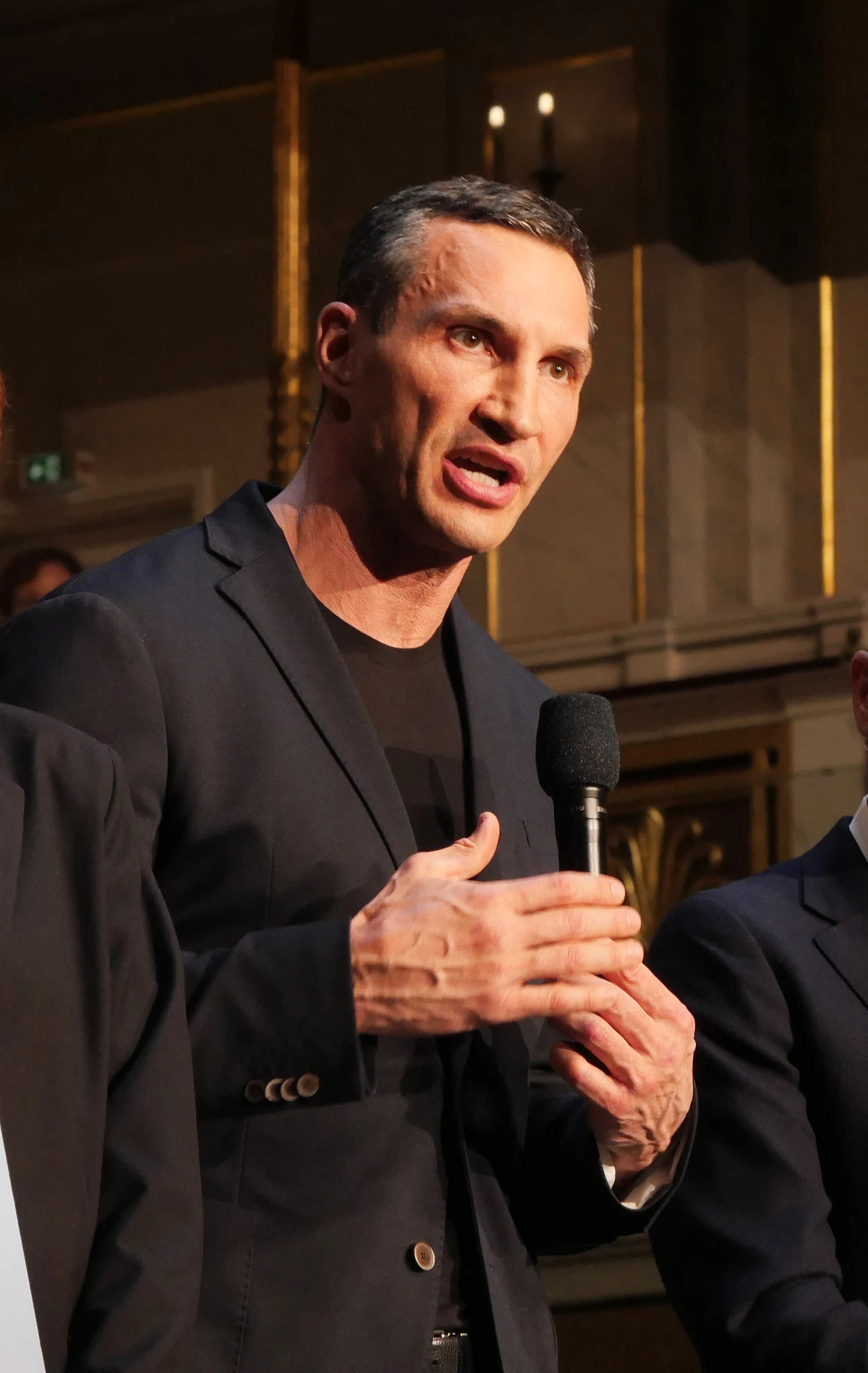 Mężczyzna w garniturze trzyma mikrofon i gestykuluje podczas przemówienia, w tle eleganckie wnętrze z elementami dekoracyjnymi.