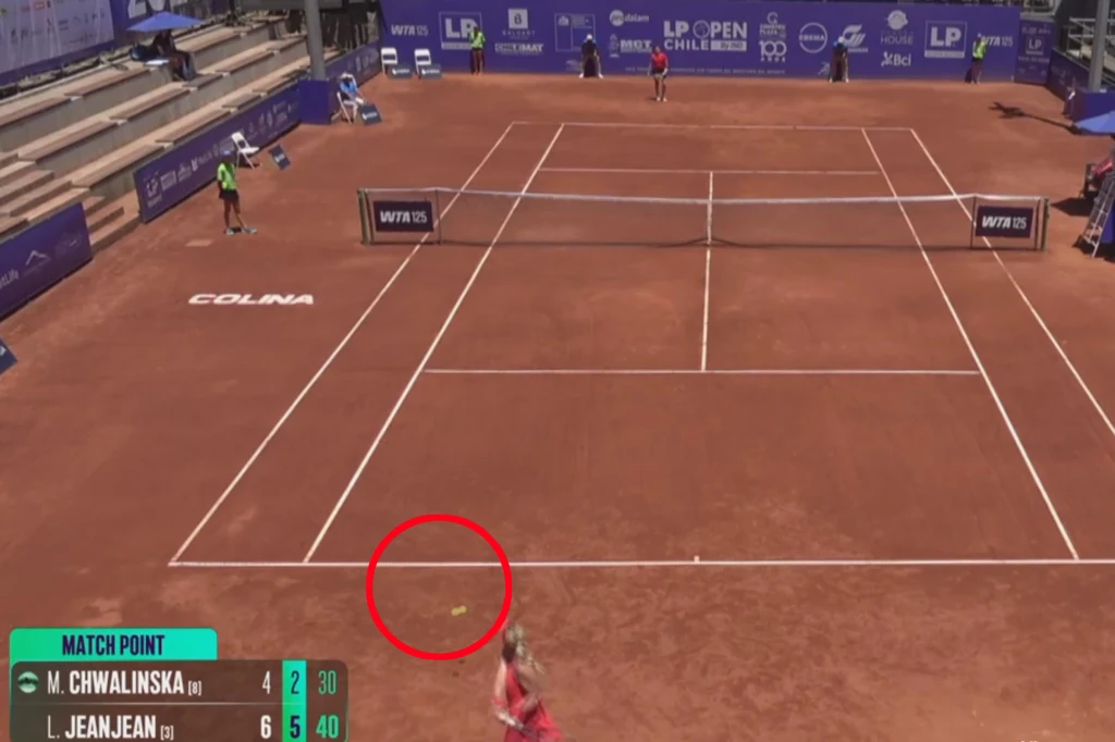 Taka Maja Chwalińska pomyliła się przy piłce meczowej dla rywalki Kort tenisowy ziemny podczas rozgrywki, w centrum widoczna czerwona linia oraz piłka tenisowa zaznaczona na żółto, jedna z zawodniczek na pierwszym planie, plansze reklamowe oraz wynik meczu w lewym dolnym rogu.