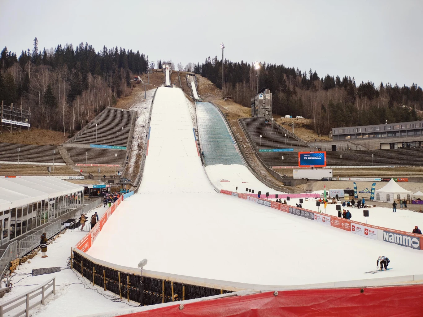 Skocznia narciarska otoczona trybunami i lasem, przygotowana do zawodów. Po bokach widoczne namioty i stanowiska techniczne oraz nieliczni pracownicy na śniegu. W tle wzgórze porośnięte drzewami iglastymi.