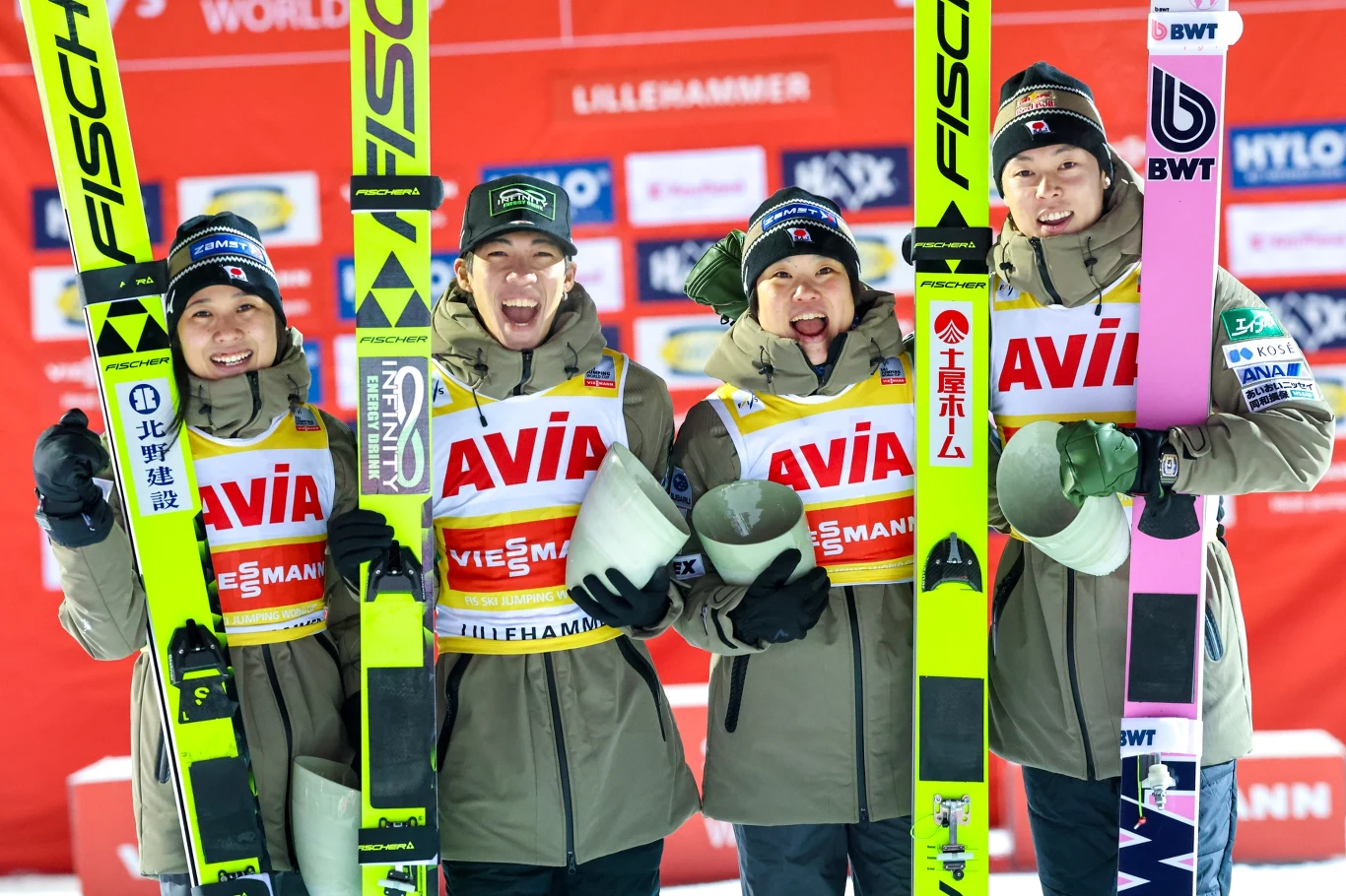Czterech skoczków narciarskich w kombinezonach drużynowych stoi razem, trzymając swoje narty oraz maskotki. W tle widoczne czerwone banery z nazwami sponsorów oraz napisem Lillehammer.
