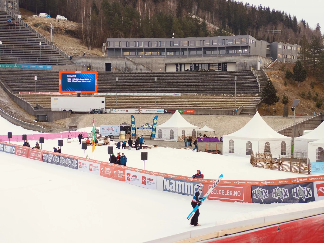 Skocznia narciarska z otaczającymi ją namiotami i banerami reklamowymi, kilka osób na śniegu, z których jedna niesie narty, a w tle widać trybuny, budynek i las.
