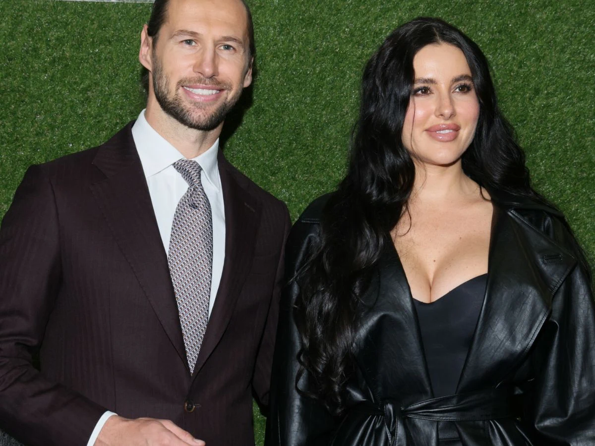 Grzegorz Krychowiak, Celia Jaunat Para elegancko ubranych osób stoi na tle zielonej, sztucznej trawy. Mężczyzna ubrany jest w bordowy garnitur i jasny krawat. Kobieta ma na sobie czarną skórzaną sukienkę z głębokim dekoltem, z długimi, rozpuszczonymi włosami.