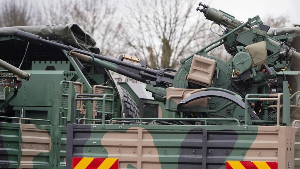 Nowoczesna armata przeciwlotnicza zamontowana na ciężarówce wojskowej, elementy sprzętu maskowane malowaniem w barwach ochronnych, w tle rozmyte drzewa.