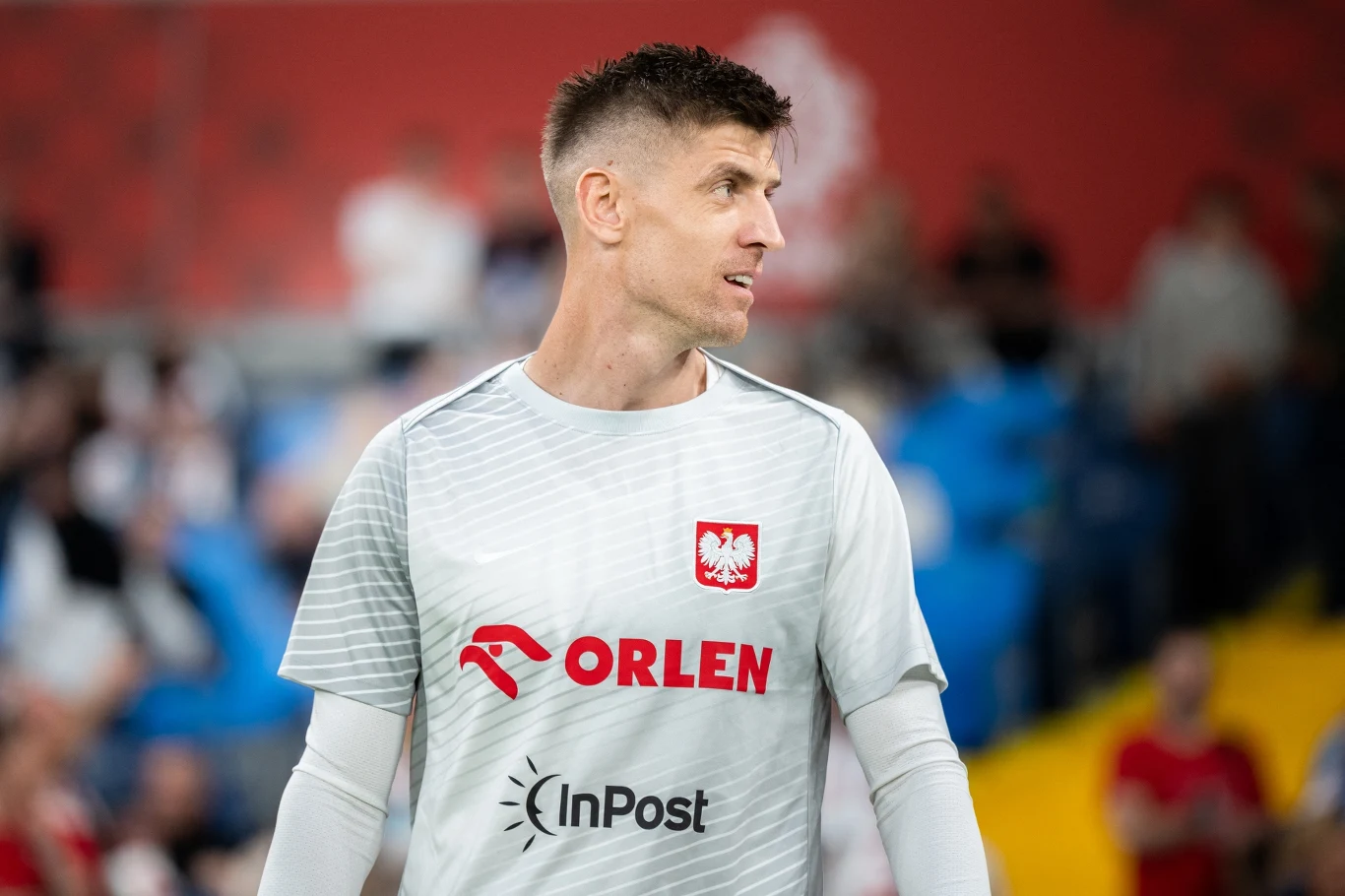 Krzysztof Piątek Piłkarz w białej koszulce treningowej z logo reprezentacji Polski oraz sponsorami, stojący na tle rozmytej publiczności i stadionu.