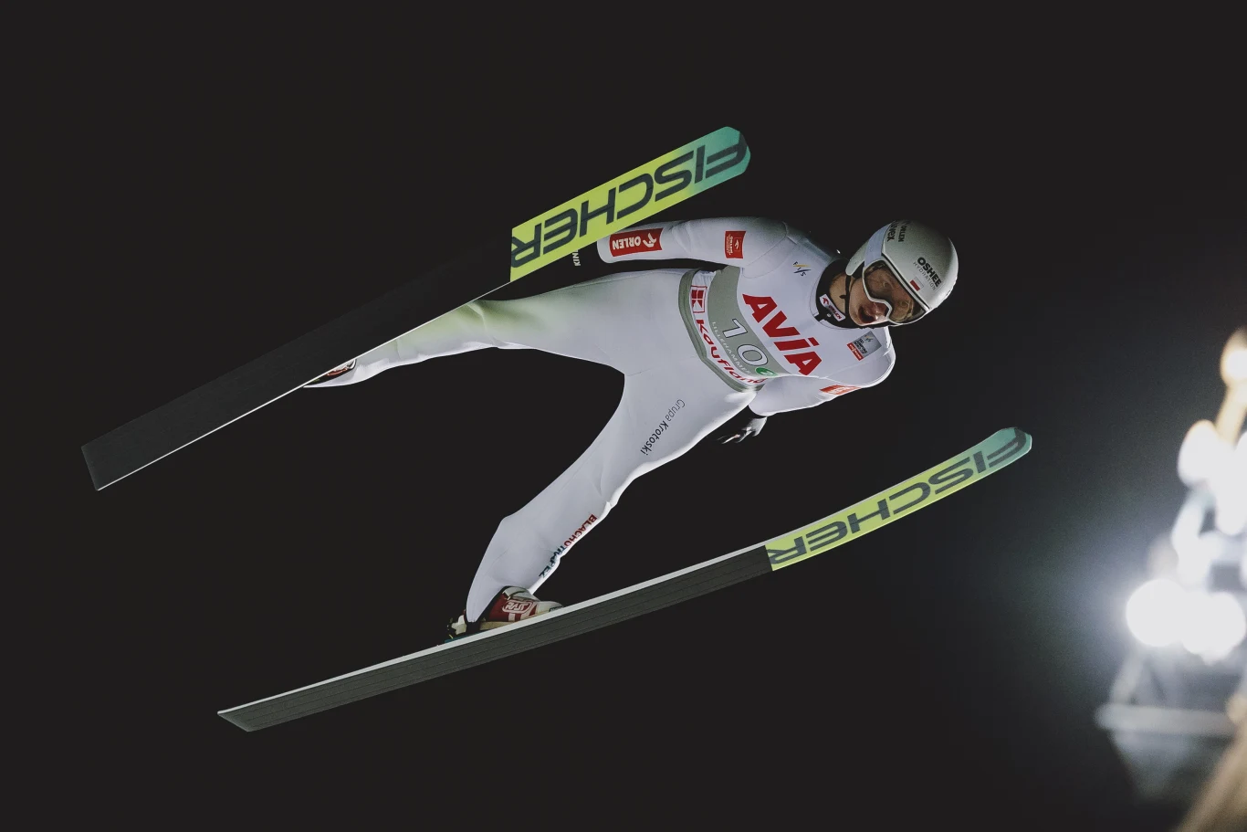 Kacper Tomasiak Zawodnik w białym kombinezonie startowym i kasku leci w powietrzu podczas skoku narciarskiego na tle nocnego nieba, narty są równoległe, w tle widoczne światła stadionowe.