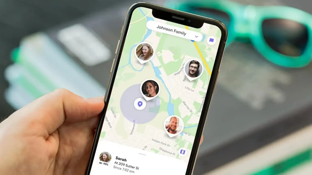 Aplikacja lokalizacyjna wyświetlająca mapę na ekranie smartfona. Na mapie widoczne są zdjęcia profilowe członków rodziny oraz zaznaczone ich aktualne lokalizacje. W tle nieostre okulary i notes.