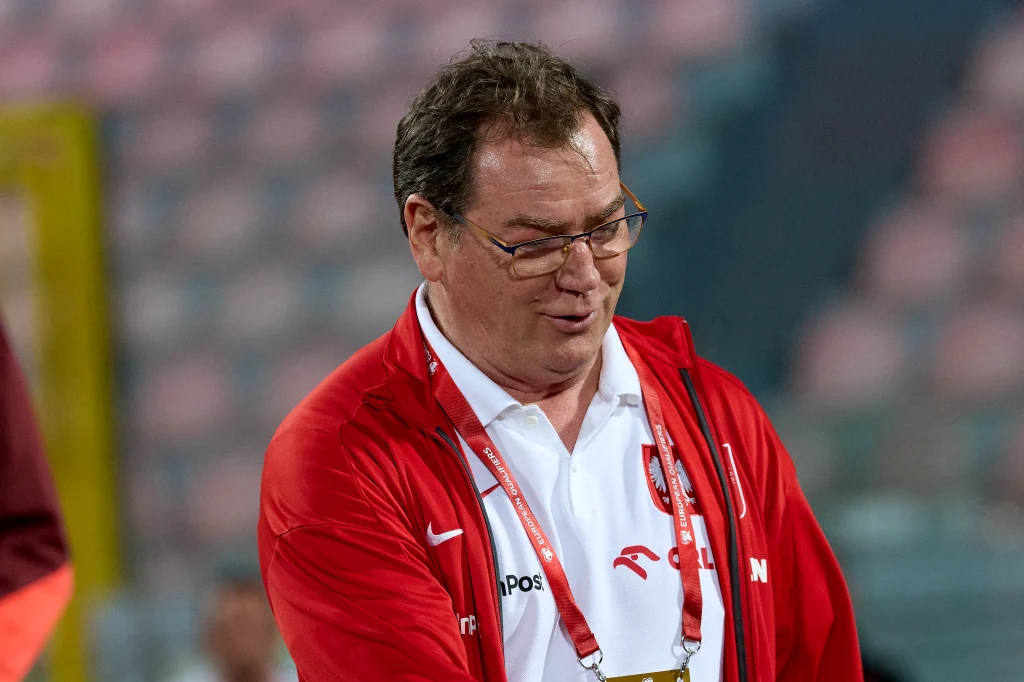 Mężczyzna w okularach ubrany w biały t-shirt oraz czerwony dres sportowy z logo drużyny, zawieszony na szyi identyfikator, rozmazane tło trybun stadionu sportowego.