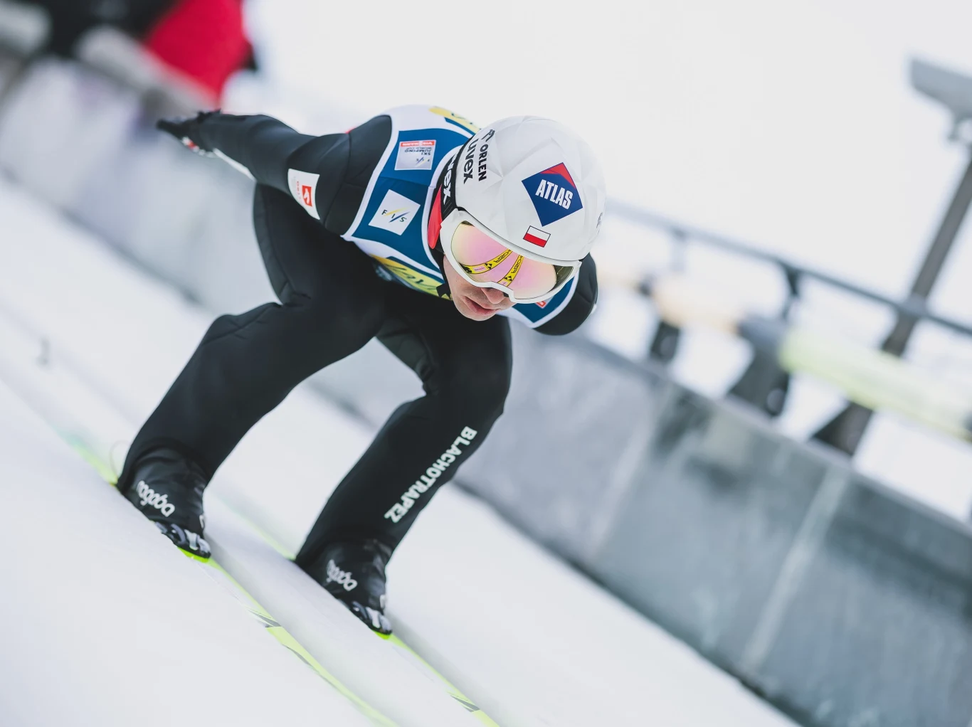 Kamil Stoch w Lillehammer Zawodnik w kombinezonie i kasku podczas skoku narciarskiego, w pozycji aerodynamicznej na rozbiegu skoczni, w tle elementy infrastruktury obiektu sportowego.