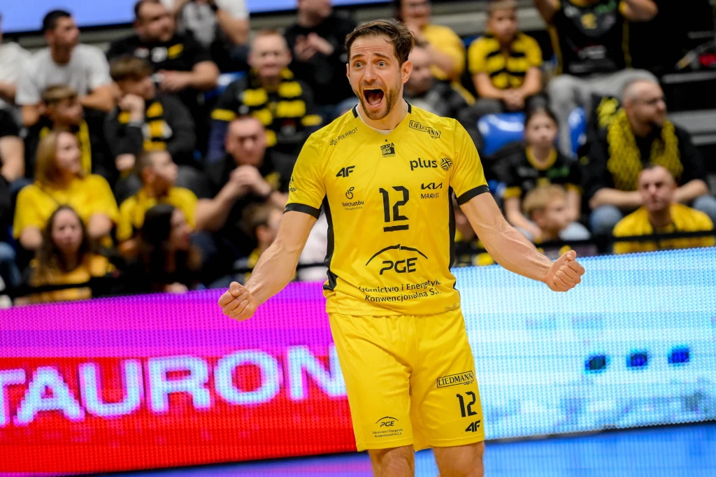 Ekspresyjny siatkarz w żółtym stroju drużynowym z szeroko otwartymi ustami, wyrażający radość lub zaskoczenie. W tle widoczni kibice w żółtych koszulkach oraz fragment elektronicznej tablicy reklamowej.