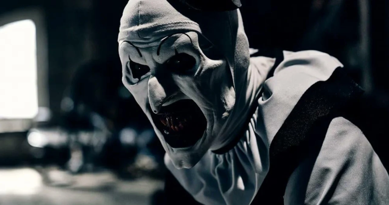 Terrifier Przerażająca postać w kostiumie klauna z białą maską i wykrzywioną, otwartą paszczą eksponującą ostre zęby, w ciemnym, mrocznym otoczeniu, podkreślającym niepokojący nastrój sceny.