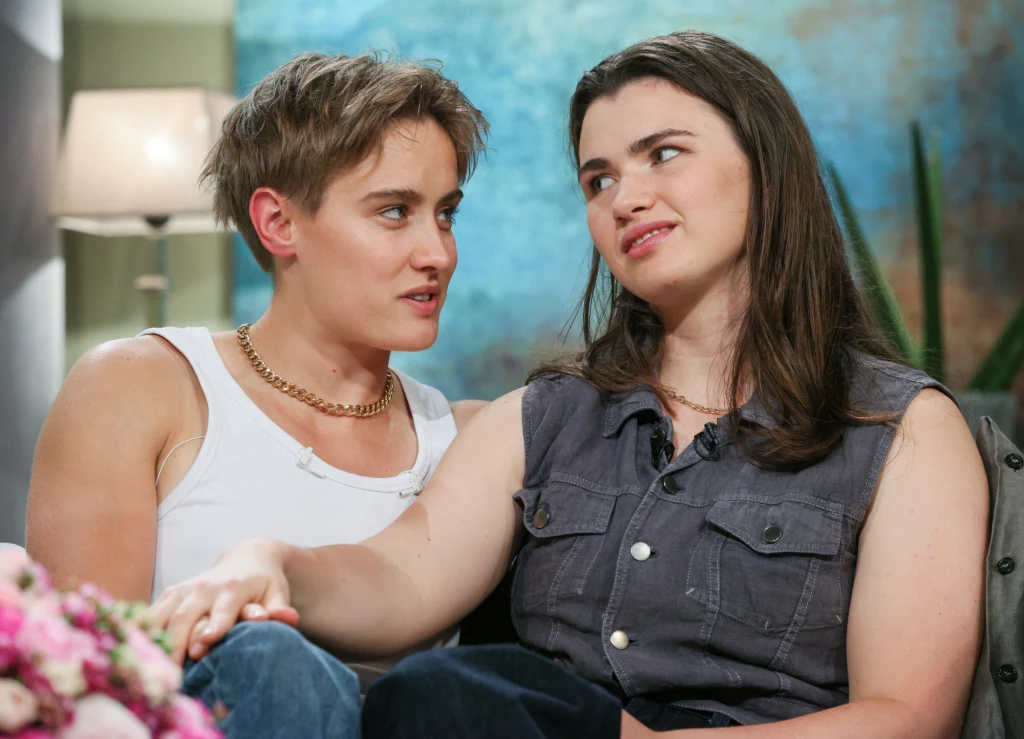 Katarzyna Zillmann, Julia Walczak Dwie osoby siedzące blisko siebie na sofie, zwracające się ku sobie z czułym i zaangażowanym spojrzeniem, jedna osoba trzyma rękę na dłoni drugiej. W tle lampa stojąca i zielona roślina dodające przytulności wnętrzu.