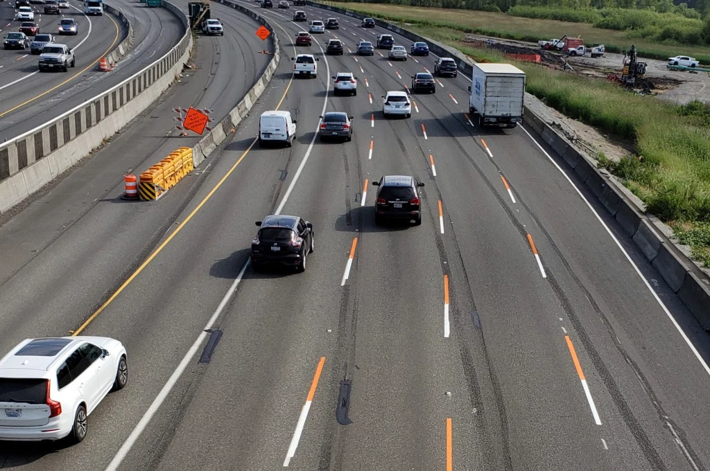 Ruchliwa autostrada z kilkoma pasami w obu kierunkach, wieloma samochodami oraz ciężarówką. Po lewej stronie widoczny betonowy separator oraz elementy prac drogowych z pomarańczowymi znakami ostrzegawczymi. Po prawej były teren budowy i zieleń.