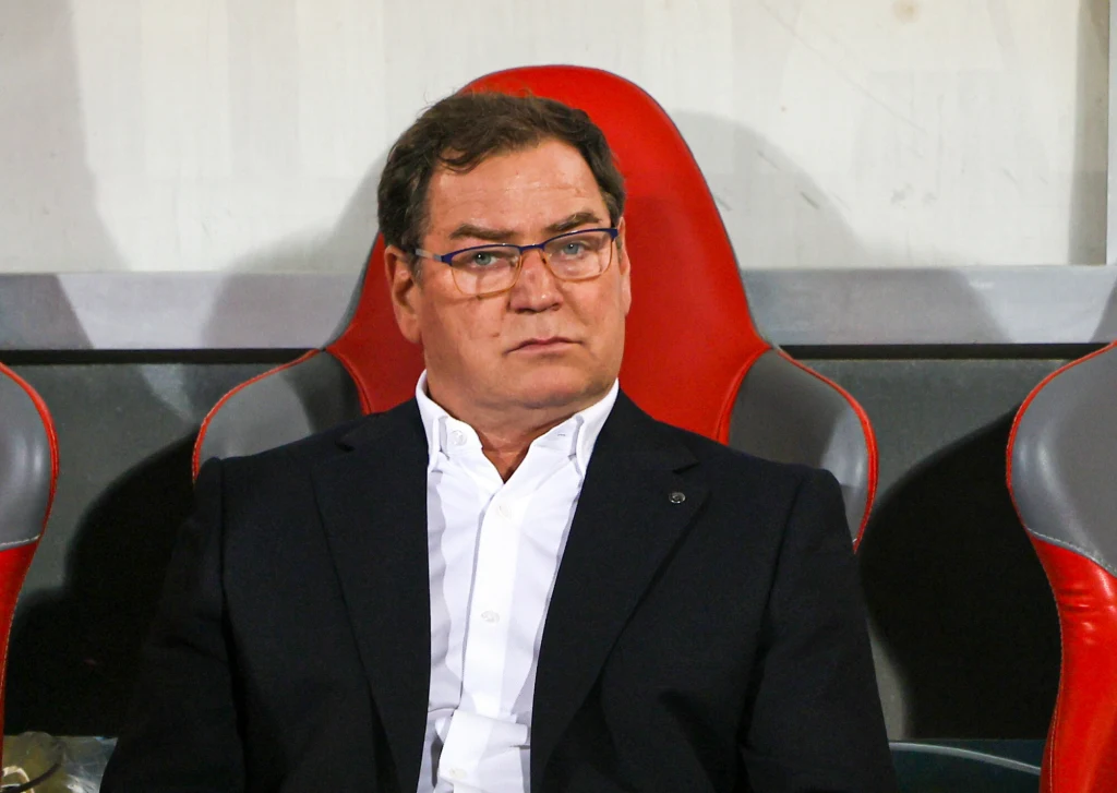 Jan Urban Mężczyzna w okularach ubrany w elegancki, ciemny garnitur i białą koszulę siedzi na stadionowym krześle o czerwonym oparciu, w tle szare i białe elementy ściany.