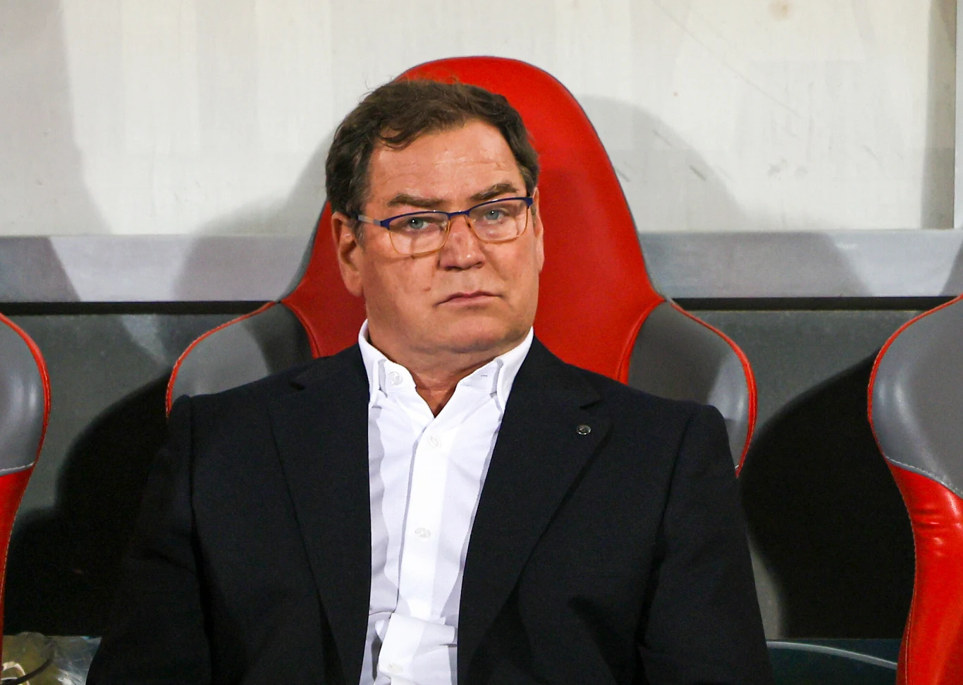 Mężczyzna w okularach ubrany w elegancki, ciemny garnitur i białą koszulę siedzi na stadionowym krześle o czerwonym oparciu, w tle szare i białe elementy ściany.