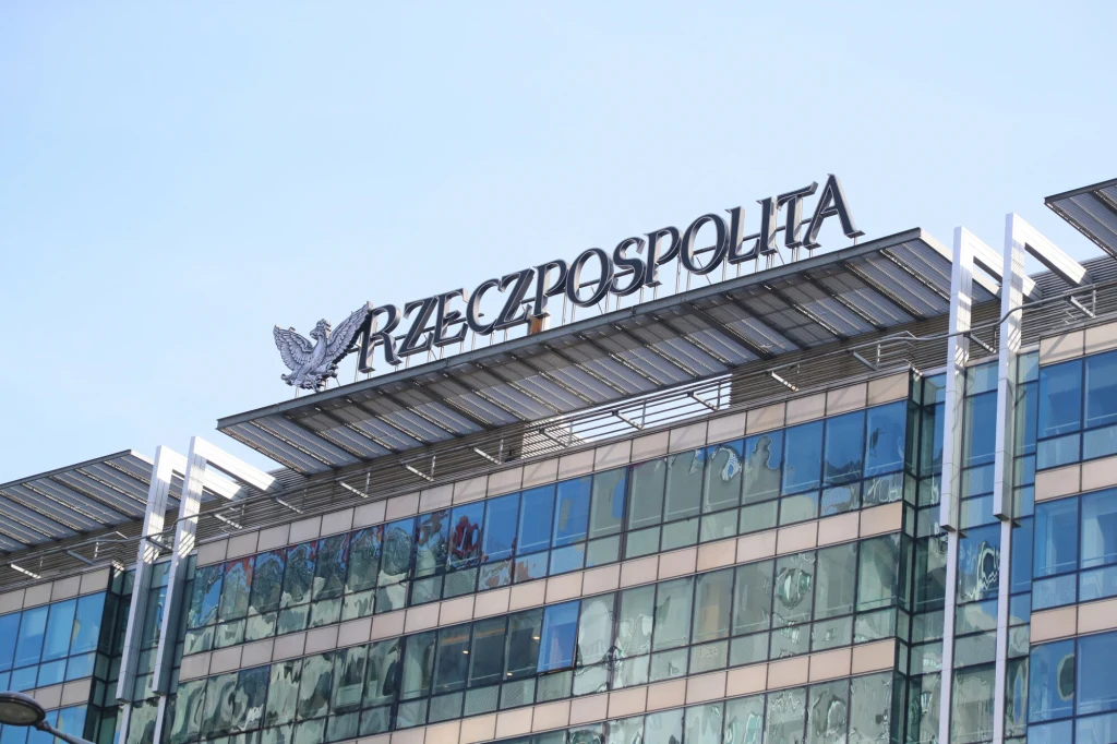 Nowoczesny szklany budynek z logo gazety Rzeczpospolita umieszczonym na dachu, z widocznym godłem orła, odbicia nieba i otoczenia na przeszklonej fasadzie.