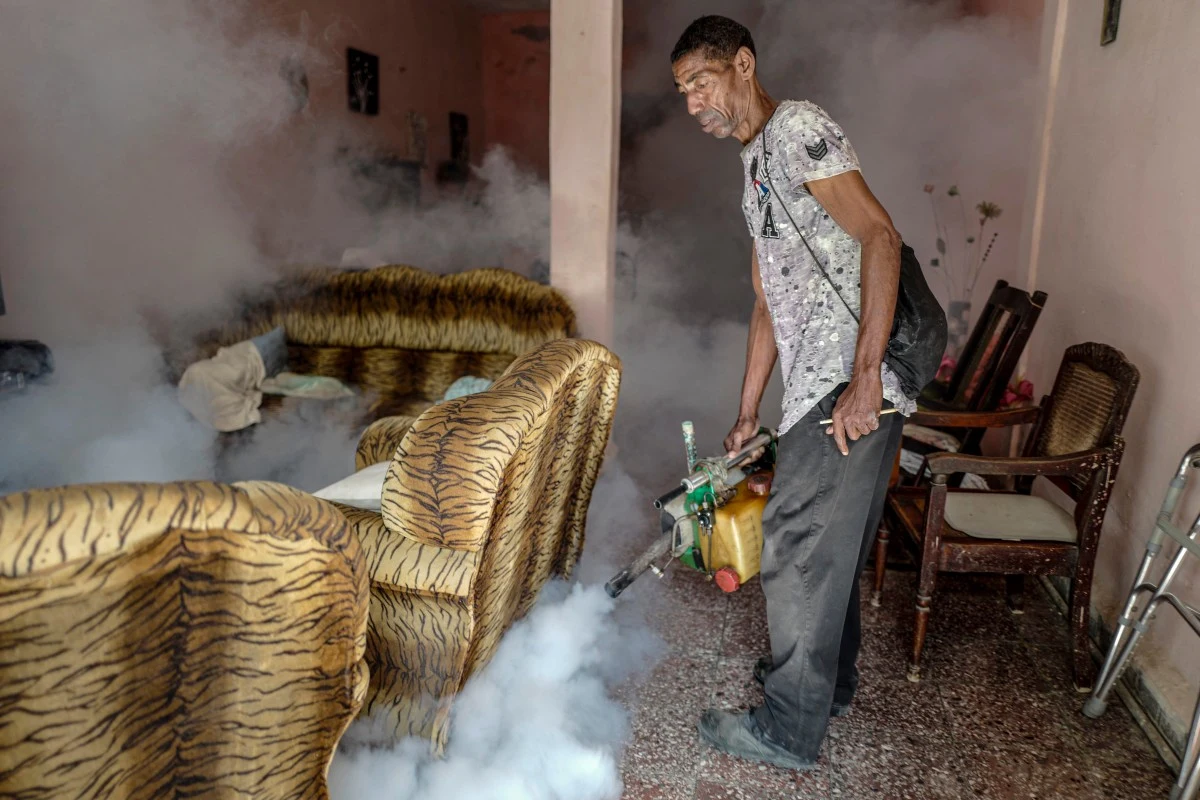 Pracownik kubańskiego Ministerstwa Zdrowia przeprowadza fumigację domu w dzielnicy Jesus Maria w Hawanie Mężczyzna używający specjalistycznego sprzętu do rozpylania dymu w salonie z fotelami o wzorze w cętki, wokół unosząca się mgła dezynfekcyjna, wnętrze mieszkalne z widocznym wyposażeniem.