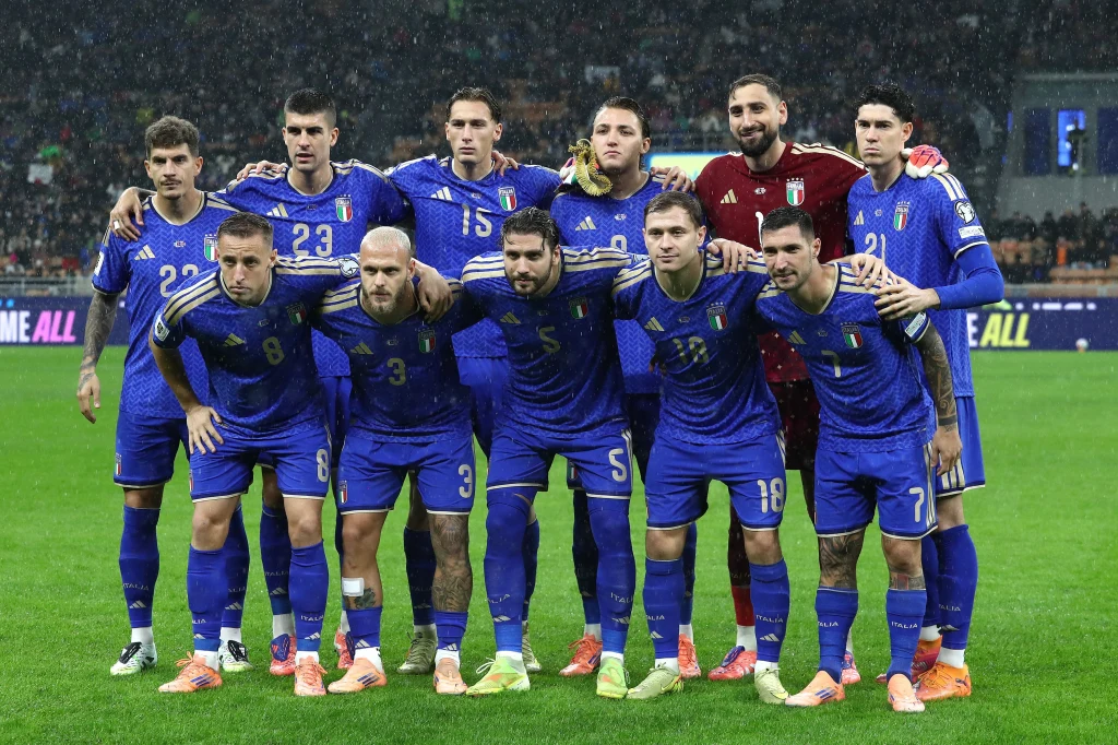 Reprezentacja Włoch Grupa jedenastu piłkarzy w niebieskich strojach drużynowych pozuje wspólnie na boisku, większość z nich stoi, a czterech kuca z przodu; w tle widoczna murawa oraz elementy stadionu sportowego.