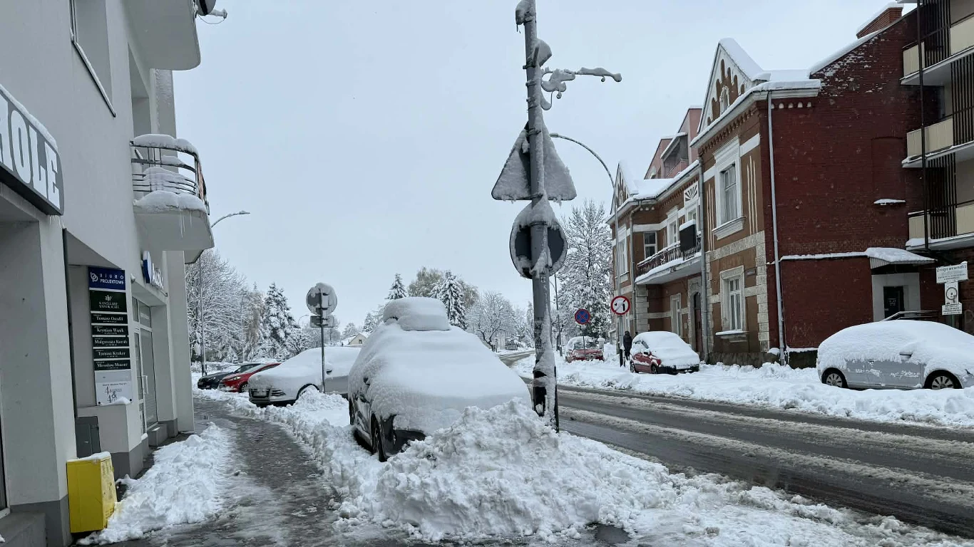 Samochody zaparkowane przy ulicy zasypane grubą warstwą śniegu, chodnik i jezdnia przykryte świeżym śniegiem, budynki mieszkalne oraz znaki drogowe częściowo zasłonięte opadami.