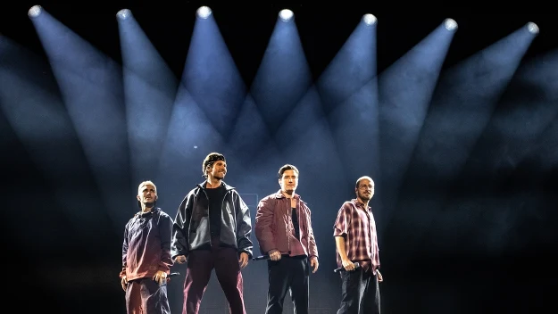 Czterech mężczyzn stoi na podwyższonej scenie, każda postać jest podkreślona przez skierowane na nich silne światła reflektorów, ubrani są w ciemne i bordowe stroje, atmosfera tworzy wrażenie oczekiwania lub rozpoczęcia występu.