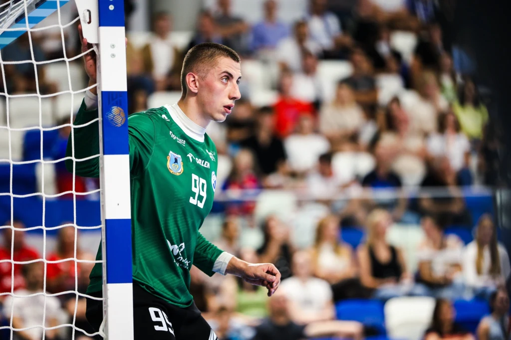 Bramkarz w zielonym stroju sportowym z numerem 99 stoi przy słupku bramki na hali sportowej, wokół widownia wypełniona kibicami.