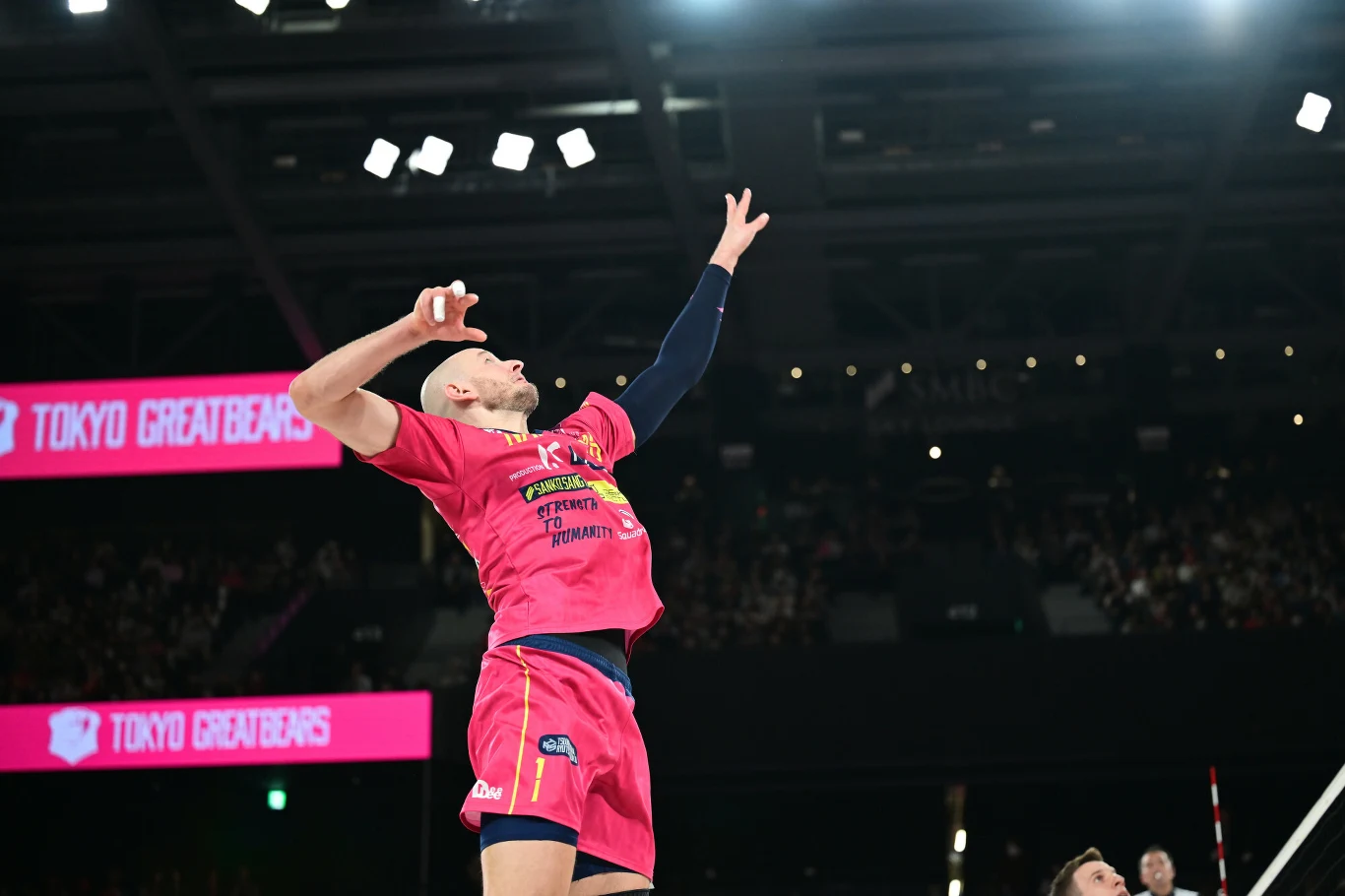 Siatkarz w różowym stroju wykonuje zagrywkę podczas meczu na hali sportowej, w tle widoczni kibice oraz jasne oświetlenie, po lewej stronie ekranu napis Tokyo Greatbears.