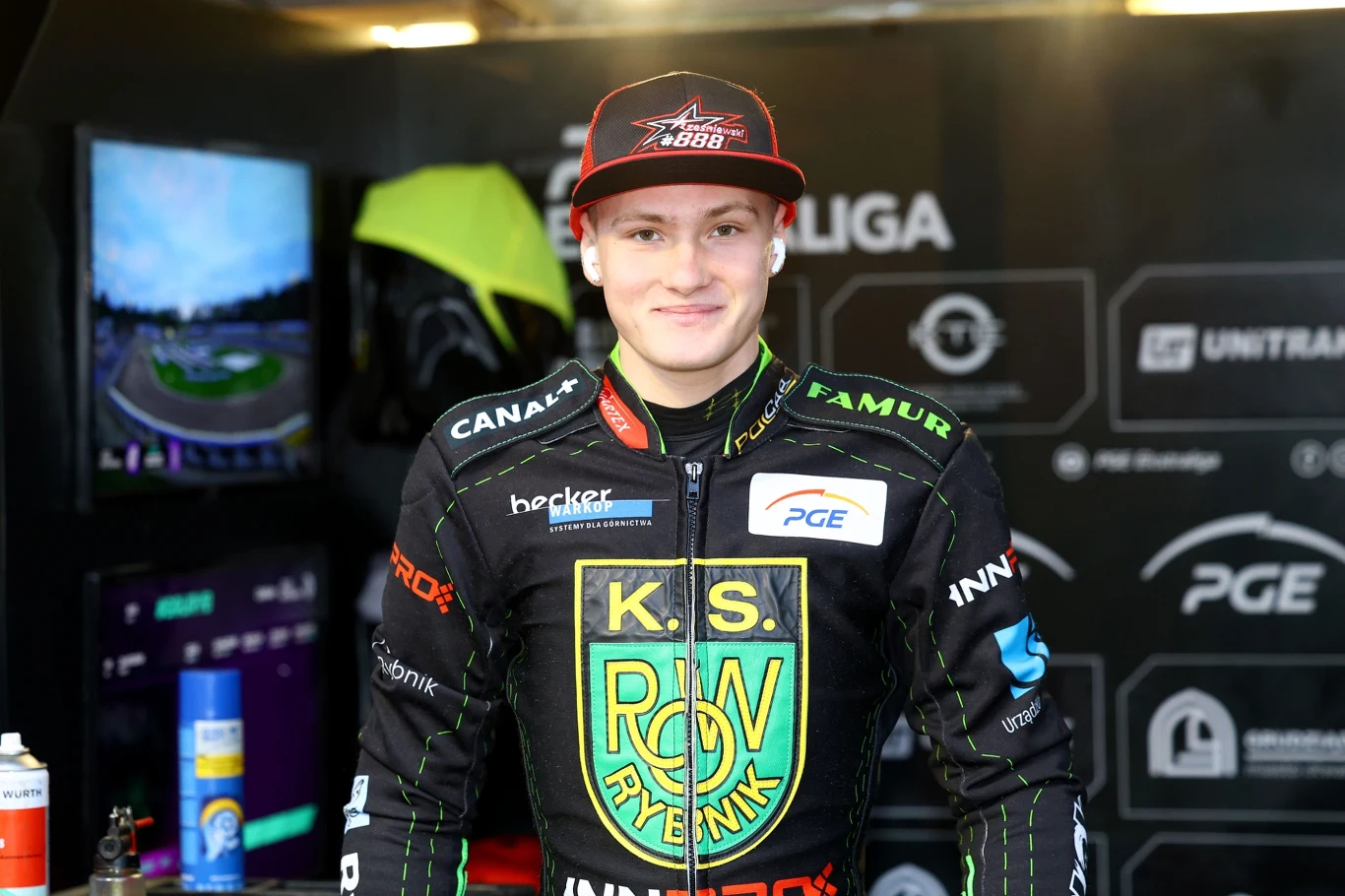 Paweł Trześniewski w barwach ROW-u Rybnik. Żużlowiec w stroju sportowym z logo K.S. ROW Rybnik stoi w boksie drużynowym, w tle widoczne elementy wyposażenia i logotypy sponsorów na ścianie.