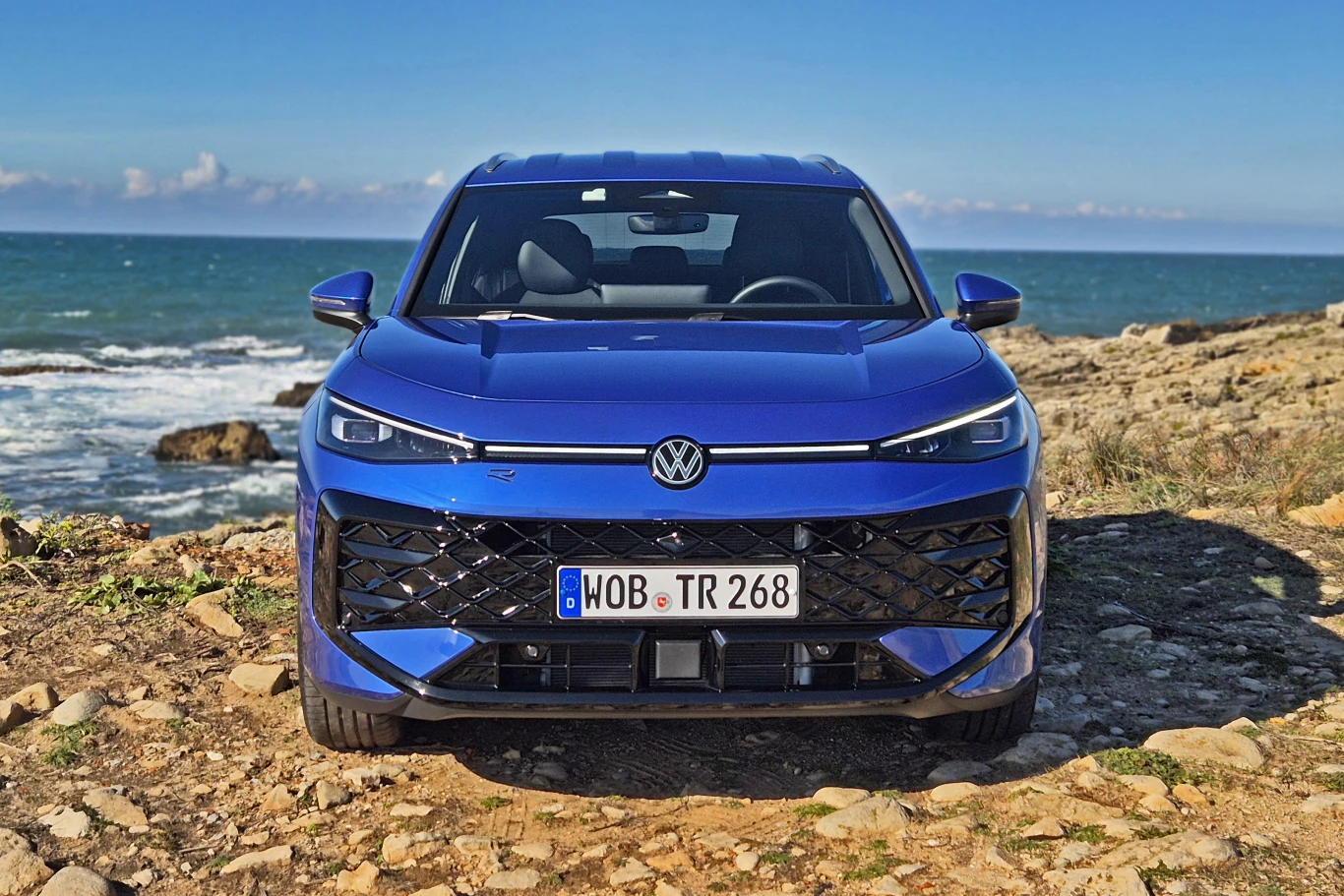 T-Roc najlepiej prezentuje się w odmianie R-Line Nowoczesny niebieski samochód osobowy marki Volkswagen ustawiony na kamienistym wybrzeżu z widokiem na morze, w tle lazurowa woda i niebo z lekkim zachmurzeniem.