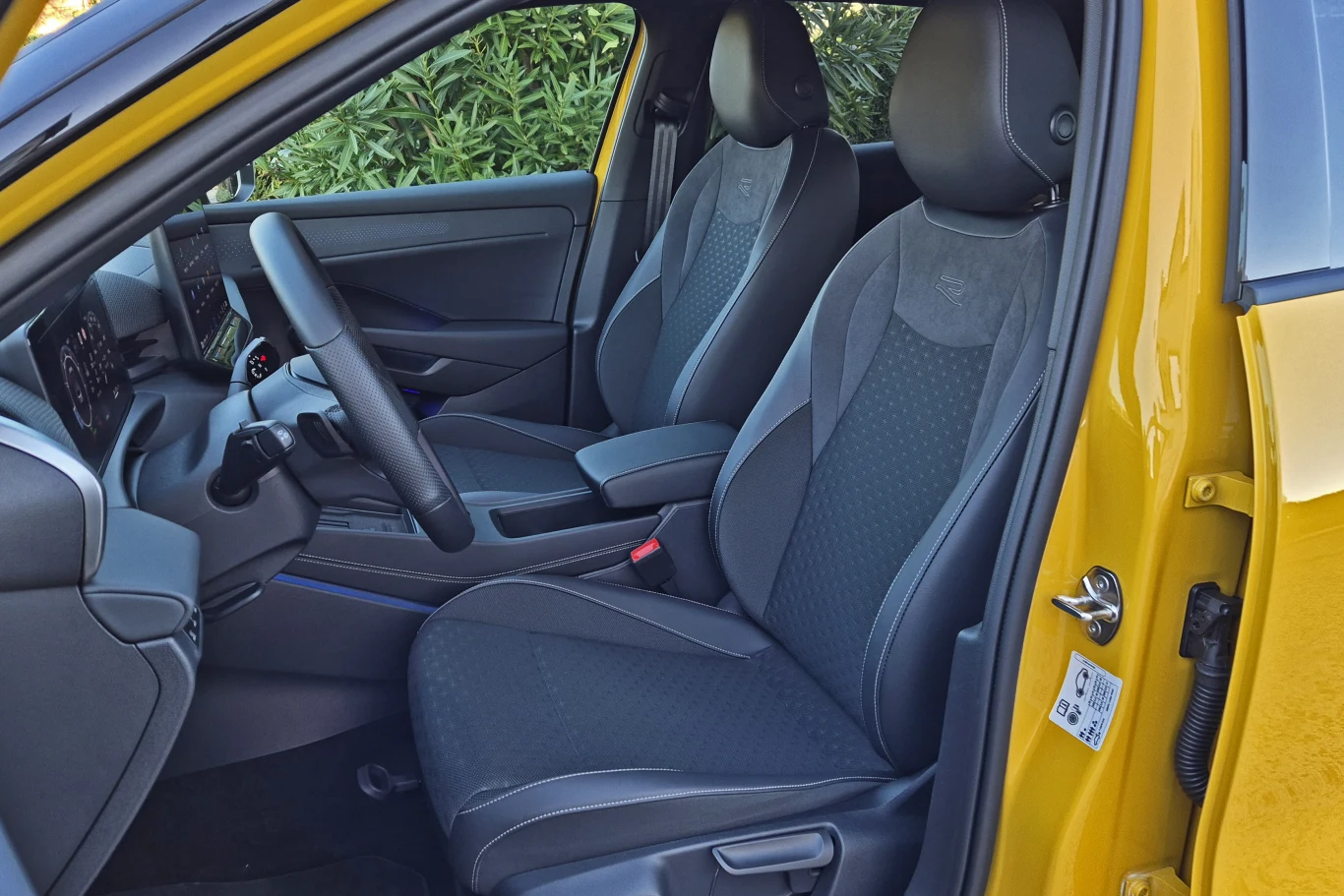Sportowe fotele w wersji R-Line są wygodne i zapewniają niezłe trzymanie boczne Nowoczesne wnętrze samochodu z żółtymi drzwiami, czarne przednie siedzenia ze sportowym wykończeniem, widoczna kierownica, deska rozdzielcza oraz fragment konsoli centralnej, całość otoczona zielenią z zewnątrz.