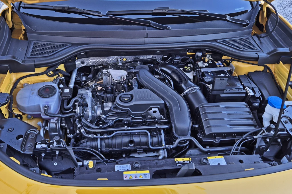 Volkswagen T-Roc początkowy dostępny jest z silnikiem 1.5 eTSI o mocy 116 oraz 150 KM Silnik samochodowy z widocznymi elementami mechanicznymi, układem przewodów, akumulatorem oraz zbiornikiem płynu chłodniczego pod maską żółtego pojazdu.