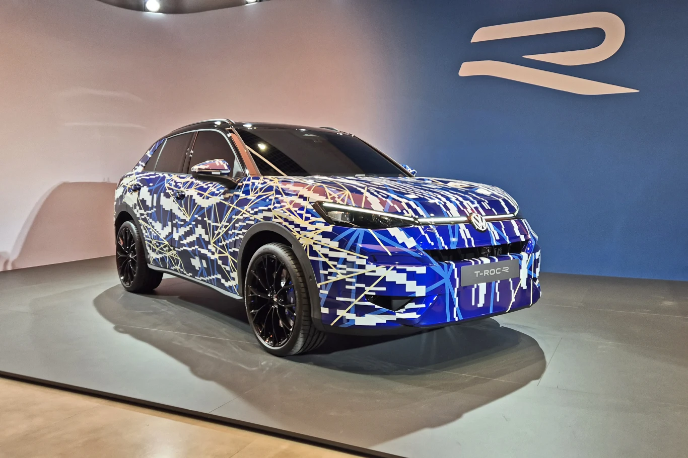Volkswagen T-Roc R jeszcze nie został oficjalnie zaprezentowany Nowoczesny samochód pokryty wzorzystym niebiesko-białym maskowaniem stoi na ekspozycji w jasnym pomieszczeniu. W tle duże logo producenta na niebieskiej ścianie.