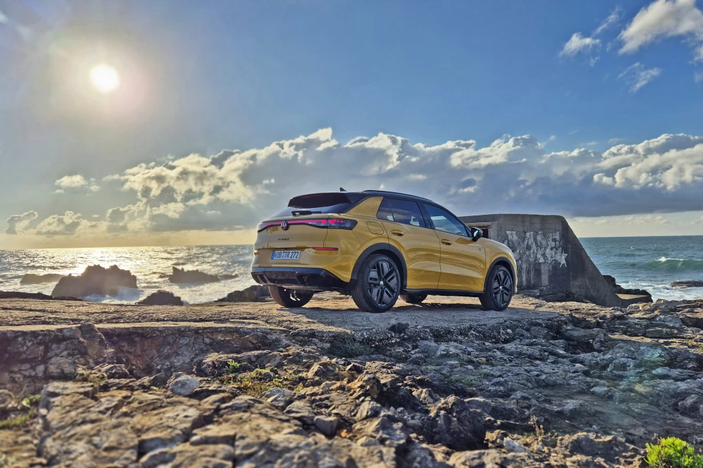 Volkswagen T-Roc dostępny jest w kilku wyrazistych kolorach oraz z lakierowanym na czarno dachem Jasnożółty samochód typu SUV stoi na skalistym brzegu w pobliżu betonowej budowli, w tle rozciąga się morze i niebo z zachodzącym słońcem oraz rozproszonymi chmurami.