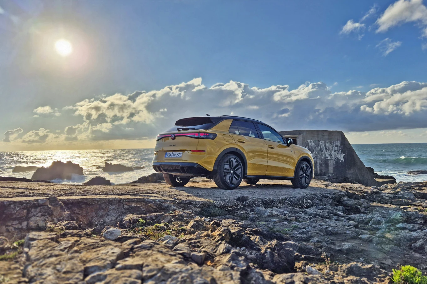 Volkswagen T-Roc dostępny jest w kilku wyrazistych kolorach oraz z lakierowanym na czarno dachem Jasnożółty samochód typu SUV stoi na skalistym brzegu w pobliżu betonowej budowli, w tle rozciąga się morze i niebo z zachodzącym słońcem oraz rozproszonymi chmurami.