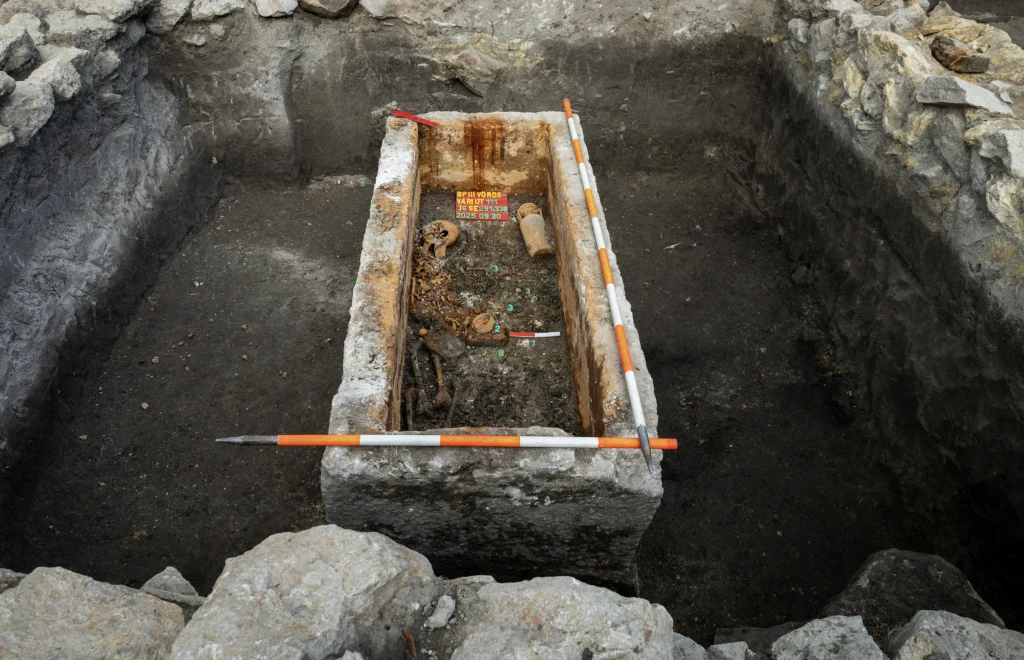 Kamienna trumna z odkrytymi szczątkami ludzkimi i metalowymi przedmiotami, umieszczona w wykopanym na ten cel grobie, otoczona przez warstwy ziemi oraz kamienne ściany wykopu. Wewnątrz widoczne narzędzia pomiarowe archeologów.