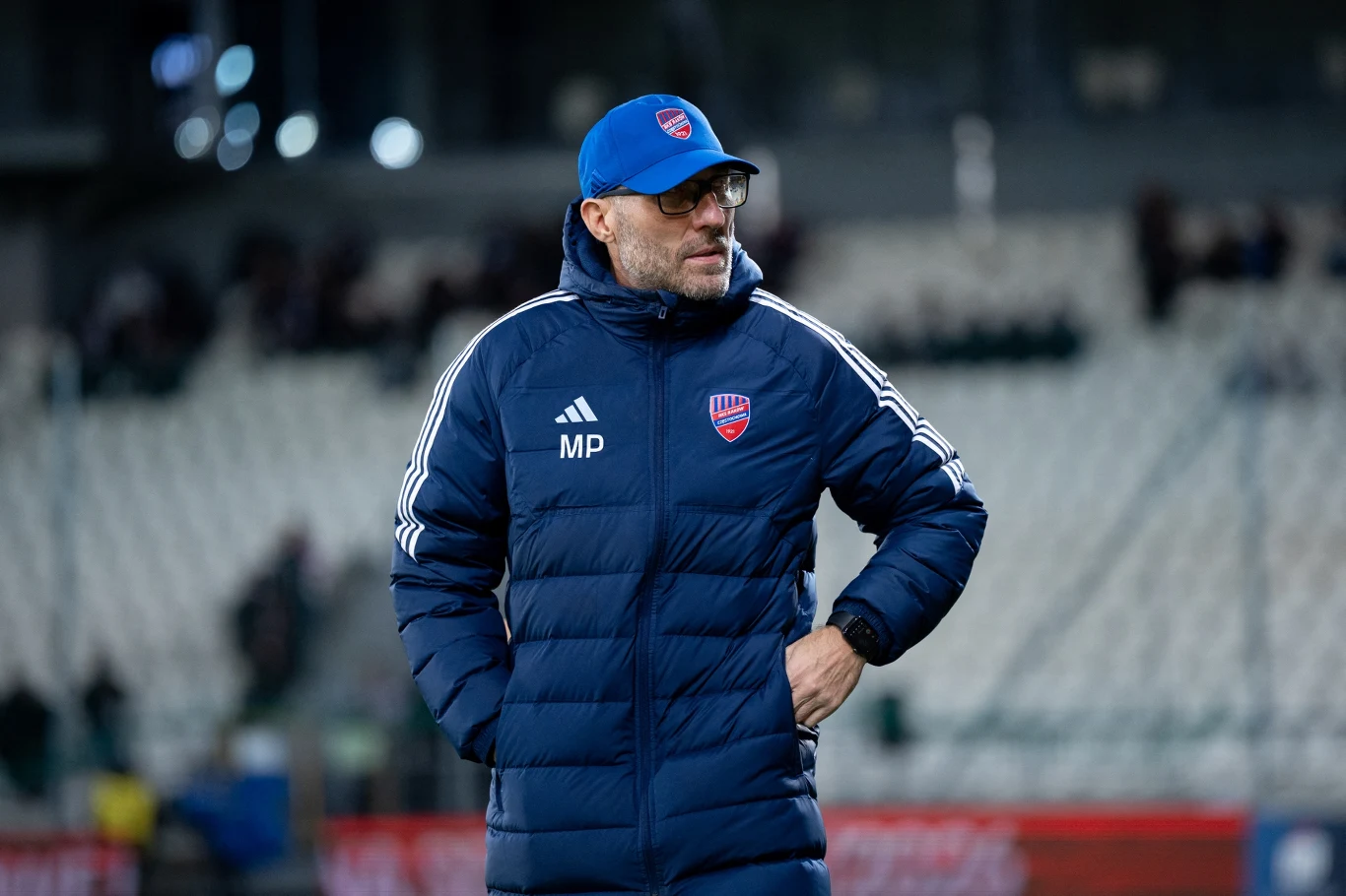 Trener drużyny piłkarskiej w niebieskiej kurtce z naszywką klubową i czapką stoi na stadionie. W tle widać zamazane trybuny oraz niewyraźnych kibiców.