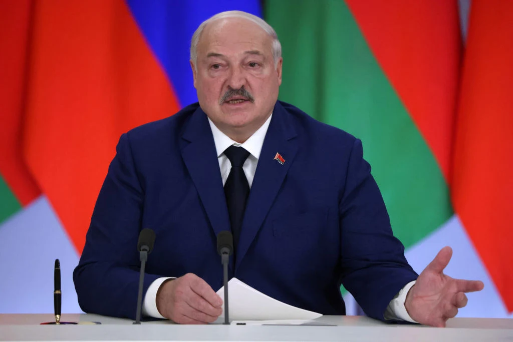 Prezydent Białorusi Alaksandr Łukaszenko Mężczyzna w granatowym garniturze siedzący przy stole z mikrofonami, trzymający dokument i gestykulujący ręką, w tle widoczne są wielobarwne flagi państwowe.