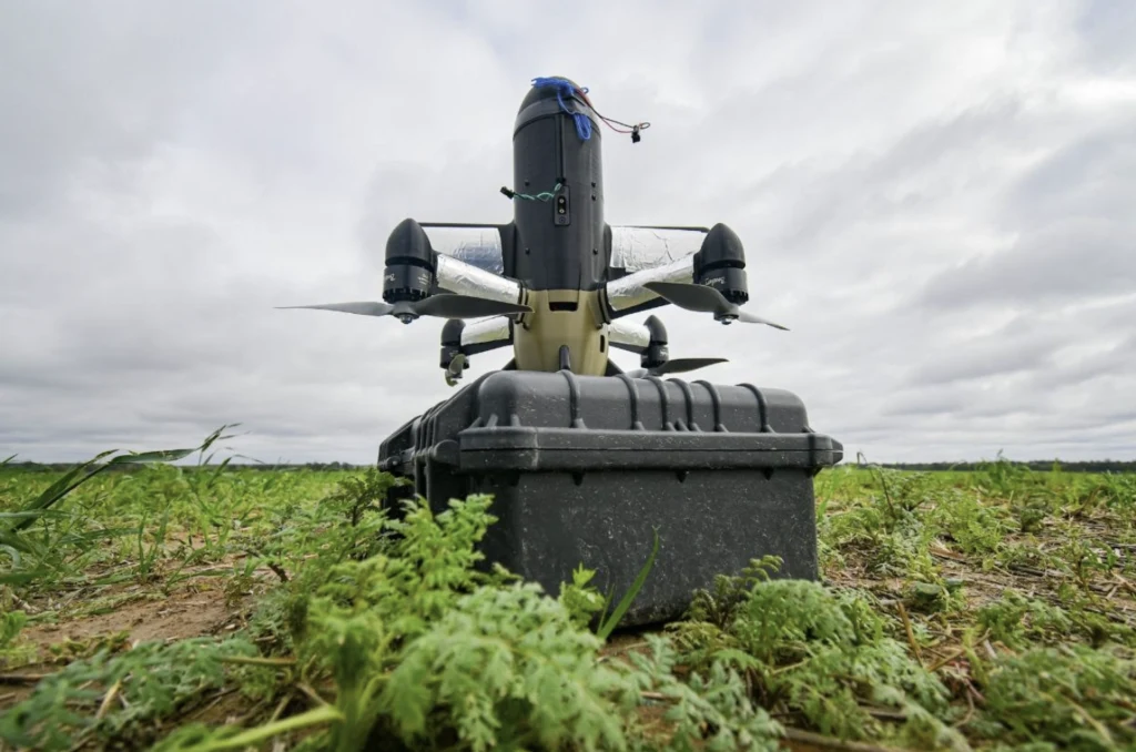 Dron stojący na czarnej walizce pośród zielonych roślin na otwartym polu, pod pochmurnym niebem.