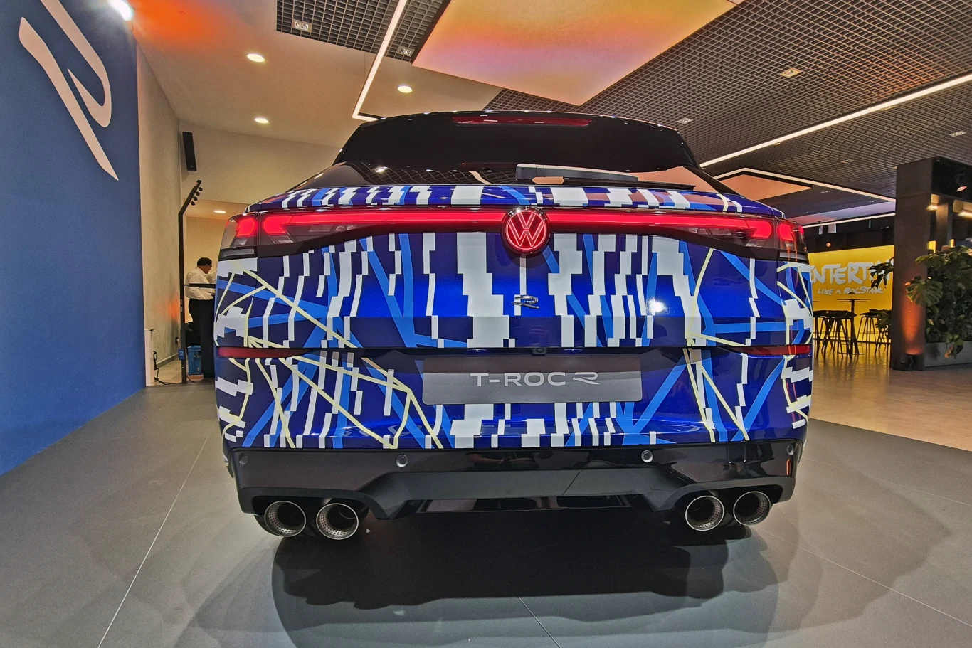 Charakterystyczny poczwórny wydech T-Roca R najpewniej nie zabrzmi zbyt rasowo z uwagi na normy i regulacje Tylny widok Volkswagena T-Roc R w nietypowym malowaniu kamuflażowym z wyraźnie widocznymi czterema końcówkami wydechu oraz nowoczesnym oświetleniem LED. Wnętrze salonu samochodowego z jasnym oświetleniem, na ścianie widoczne logo producenta.